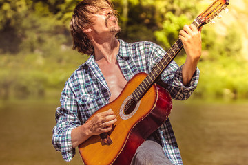 Rock guitarist in an open shirt enthusiastically plays the old guitar in nature. Summer, sunny day....
