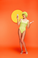 Shocked woman in hat with decorative flowers looking at smartphone with yellow circle behind on orange