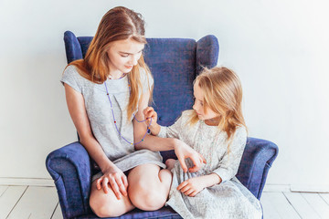 Obraz na płótnie Canvas Stay Home Stay Safe. Two happy kids sitting on cozy blue chair relaxing playing in white living room indoors. Little girl playing with teenage girl showing her love care. Sisters having fun at home.