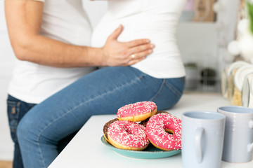 Obraz na płótnie Canvas Man holding belly of his pregnant wife, sitting on the table in the kitchen with donuts in front