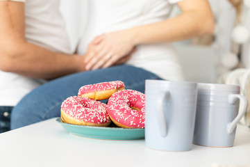 Man holding belly of his pregnant wife, sitting on the table in the kitchen with donuts in front