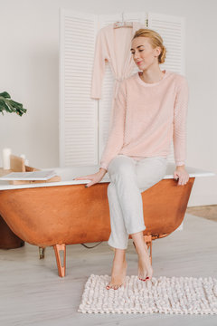 A young blonde is sitting in her pajamas, barefoot, on the edge of the bathroom, smiling thoughtfully