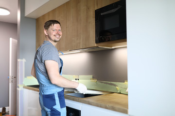 Happy man in work clothes sets hob in kitchen. Installation an induction hob above oven. Work in...