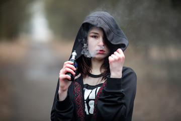 Vape teenager. Young pretty caucasian brunette girl with problem skin in the hood smoking an electronic cigarette on the street in the spring. Deadly bad habit.