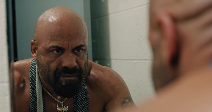 Bald, tan, muscular, bearded man looks up at himself in a dirty bathroom mirror.