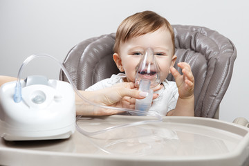 Mother using nebulizer for cute little baby boy sitting on the high chair. Health care concept.