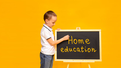 Cute schoolboy in a white T-shirt on a yellow background writes with his hand on the blackboard. Banner. Place for text. School time. Home education. Quarantine. Time to school. Home education.