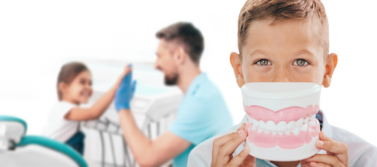Pediatric dentistry, children teeth treatment. Boy with a large model of smile in front of the face, against the background of a dentist and a girl