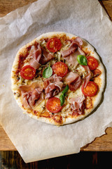 Fresh baking homemade pizza napolitana with prosciutto ham, cheese, tomatoes, basil on baking paper over wooden table background. Home baking or delivered fast food. Flat lay, space.