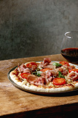 Fresh baking homemade pizza napolitana with prosciutto ham, cheese, tomatoes, basil on plate, glass of red wine over wooden table background. Home baking or delivered fast food.