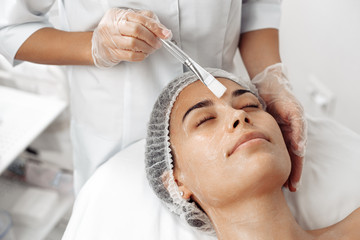 Cosmetologist applying cream on female face with brush in cosmetology salon.