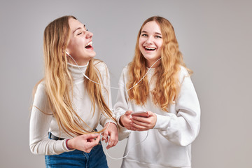 Young women girls listening to music together streaming content having fun watching video enjoying video chat talking with friends making gestures faces using smartphone earphones headphones