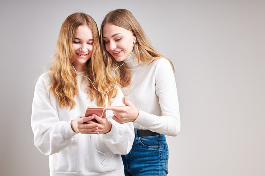 Young women girls listening to music together streaming content having fun watching video enjoying video chat talking with friends making gestures faces using smartphone earphones headphones
