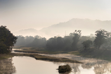 A beautiful view of a river with its bank covered with dense evergreen rainforest mountains captured during an early foggy morning at Kaziranga National Park, Assam, Northeast, India. 