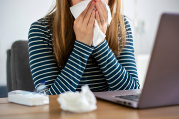 Sick woman working on laptop at home, covering nose and mouth when coughing and sneezing in tissue, ill freelance woman with cold. Coronavirus COVID-19 reducing of risk of spreading the infection.
