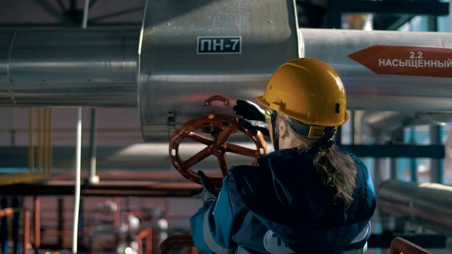 Woman worker in helmet at factory rotates red valve of big pipe
