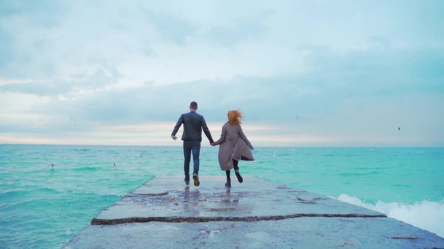 young loving couple running on the pier. holding hands, cold weather, rain, wind, birds flying, seagulls. big waves, winter or spring or fall, dark clothing, coat, seashore or ocean, slow motion. Sea