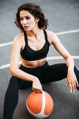 Beautiful caucasian dark hair girl in sportswear holding basketball sitting at the court outside. Energetic sexy fitness trainer wearing black leggings and sport bra during workout