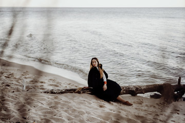 A girl in a black dress and white shirt sits on a log on a deserted seashore in cloudy weather and on a calm day.