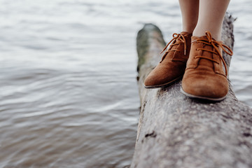A girl in brown leather boots is standing on a log on the seashore, waves with white foam are rolling on the shore against the background - Powered by Adobe