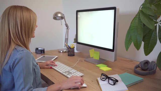 Blonde Caucasian woman works from her home office. Modern workspace with computer, tablet and notebook on a wooden table. She wears casual clothes with a denim shirt.