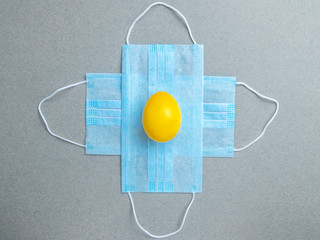One yellow easter egg lies on blue medical masks in the shape of a cross on a gray background. Flat lay. Easter holiday concept during quarantine coronavirus epidemic. Copy space