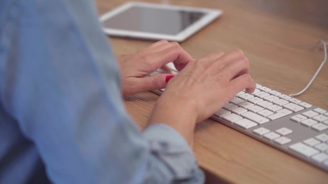 Close up of a woman's hands typing on the white computer keyboard on a wooden desk