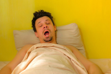 Man with hangover wakes up, yawns and rubs his eyes in bed, portrait closeup. Young man with...