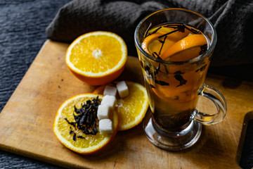 Citrus drink, tea with petals in a transparent glass mug with slices of sweet orange and refined sugar. Slicing fruit on a wooden Board with a brown napkin in a cozy dark background in a low key