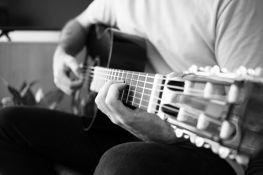 Close up of a guy playing the acoustic guitar at home, black and white photo, selective focus. Guitar player practicing guitar scales.