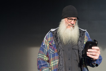 Mature bearded hipster man using phone against black wall