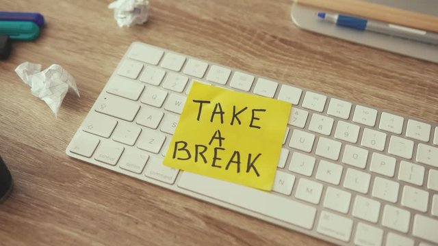 Close-up man lays a sticker note on keyboard saying Take a Break. Messy desktop workplace, lack of ideas. Concept of rest, business, motivation.