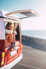 Happy adult beautiful woman enjoy travel lifestyle or summer holiday vacation with old vintage van transport sit down on the back and looking the road and ocean - people free to move and live