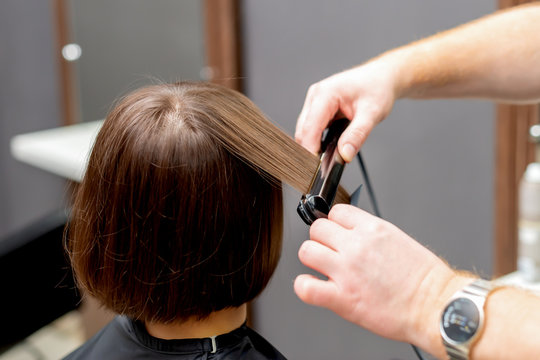 Hairdresser hands are straightening hair of woman with straightener tool in hair salon, back view.