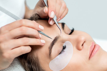 Cosmetologist hands are holding tweezers above the face of young woman during eyelash extensions procedure, close up.