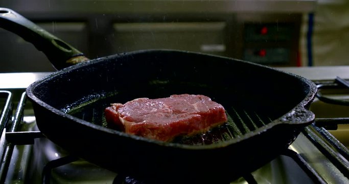 Close-up of the chef's hands in the kitchen, frying a steak in a grill pan on the stove, oil spatters flying, blue gloves on his hands.