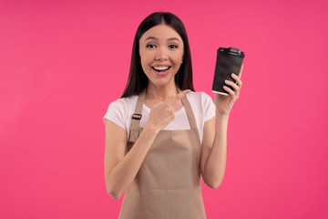 Nice stylish brunette asian girl, barista in casual tshirt and apron, points to a paper cup of tea or coffee, isolated over pink background
