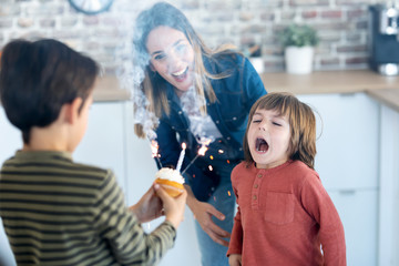 Funny boy blowing out the candles on his birthday cake while his brother is holding it at home.