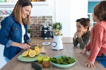 Pretty young woman cutting an apple for preparing detox beverage while her sons look her in the...