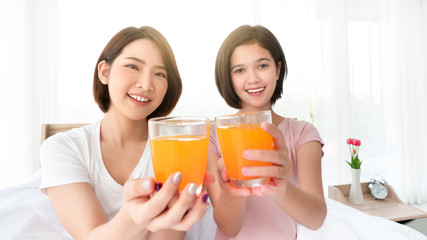 Asian young two woman smile and show closeup glass of organic orange juice beverage with nutrition and vitamin C for breakfast fresh drink within sunlight in the morning from bedroom at home