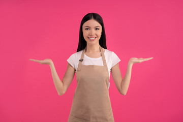 Nice stylish brunette asian girl, barista in casual tshirt and apron, holds a conditional object in her hands. free space for your advertising isolated over pink background