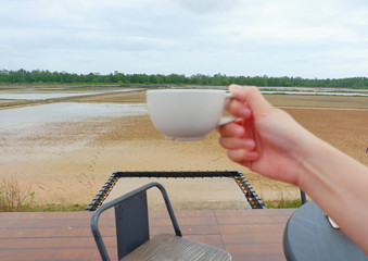 Hand women hodling white coffee cup with salt farm background. She hand keep cup for drinking coffee for relaxing on holiday time.CGirl is holding white cup in hands. Mug for woman, gift.Close up.