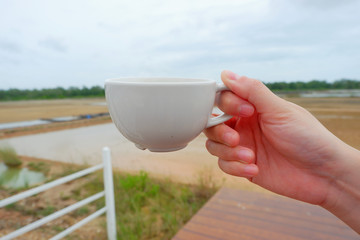 Hand women hodling white coffee cup with salt farm background. She hand keep cup for drinking coffee for relaxing on holiday time.CGirl is holding white cup in hands. Mug for woman, gift.Close up.