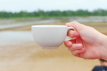 Hand women hodling white coffee cup with salt farm background. She hand keep cup for drinking coffee for relaxing on holiday time.CGirl is holding white cup in hands. Mug for woman, gift.Close up.