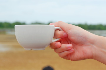 Hand women hodling white coffee cup with salt farm background. She hand keep cup for drinking coffee for relaxing on holiday time.CGirl is holding white cup in hands. Mug for woman, gift.Close up.