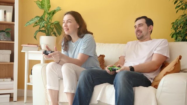 Cheerful Competitive Couple Sitting on a Couch and Playing Video Games on Console with Wireless Controllers. Yellow and White Home Interior with House Plants.