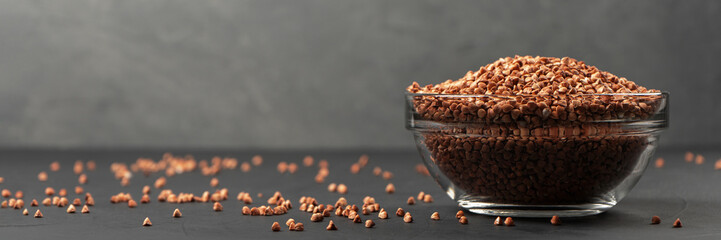 Buckwheat in a glass bowl near the copy space with spilled cereals on a black background. Panorama.