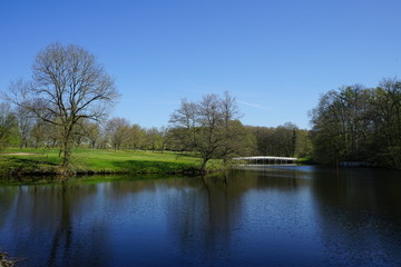 Parklandschaft mit See / Teich über den eine weiße Brücke führt im Frühling