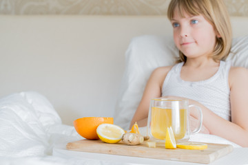 Obraz na płótnie Canvas Little sick girl in bed with cup of antipyretic drugs for colds,flu.Tea with citrus vitamin C,ginger root,lemon,orange.Wooden tray. Home self-treatment.Medical quarantine covid-19 coronavirus therapy