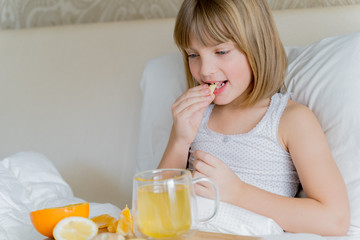 Sick girl in bed tastes ginger root piece.Cup of antipyretic drugs for colds,flu.Tea with citrus vitamin C,lemon,orange.Wooden tray.Home self-treatment.Medical quarantine therapy.Covid-19 coronavirus